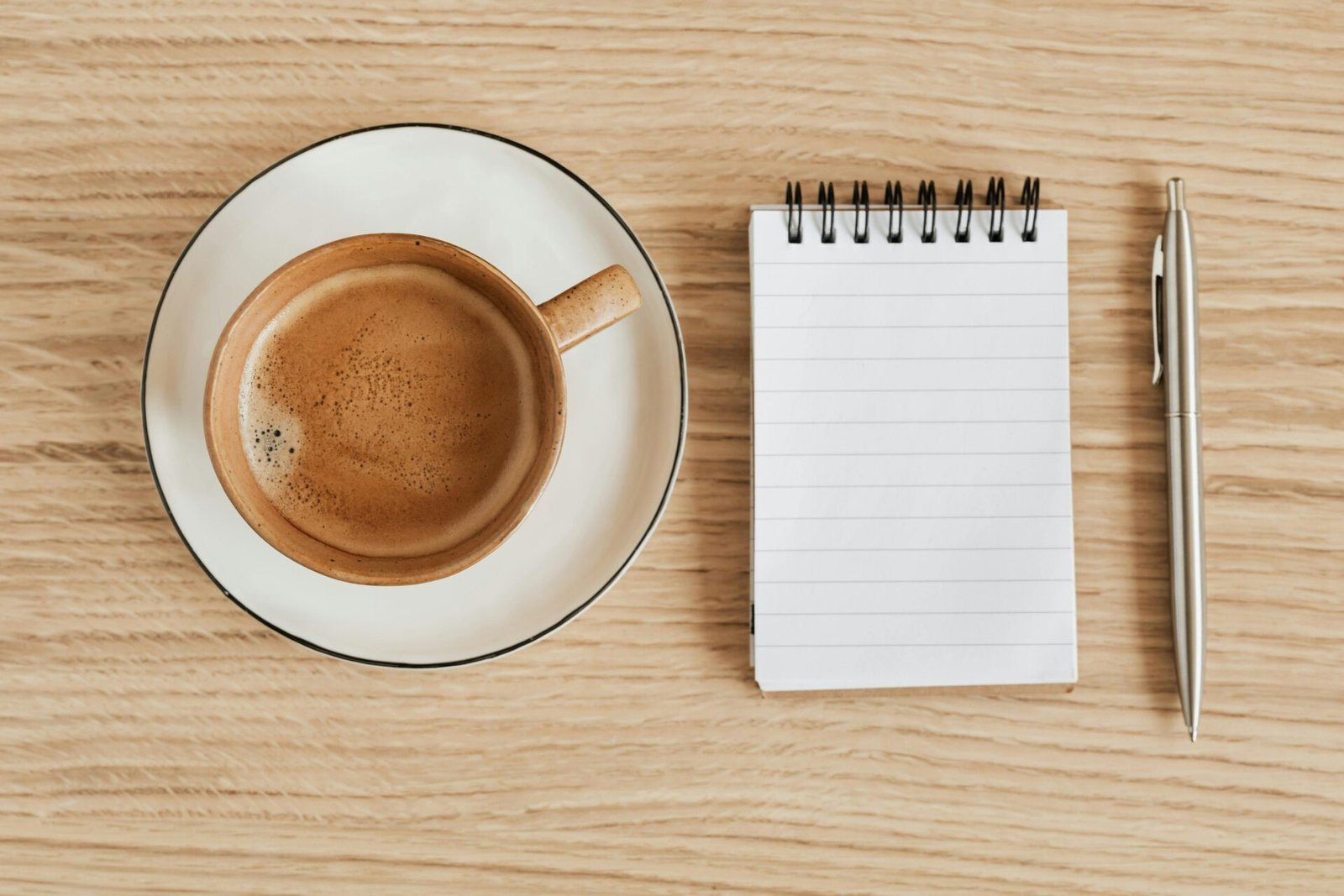 Eine Tasse Kaffee steht auf einer Untertasse neben einem leeren Spiralblock und einem silbernen Stift auf einer hellen Holzoberfläche. Der dunkle Kaffee füllt den größten Teil der Tasse und schafft so eine gemütliche und organisierte Arbeitsplatzatmosphäre.