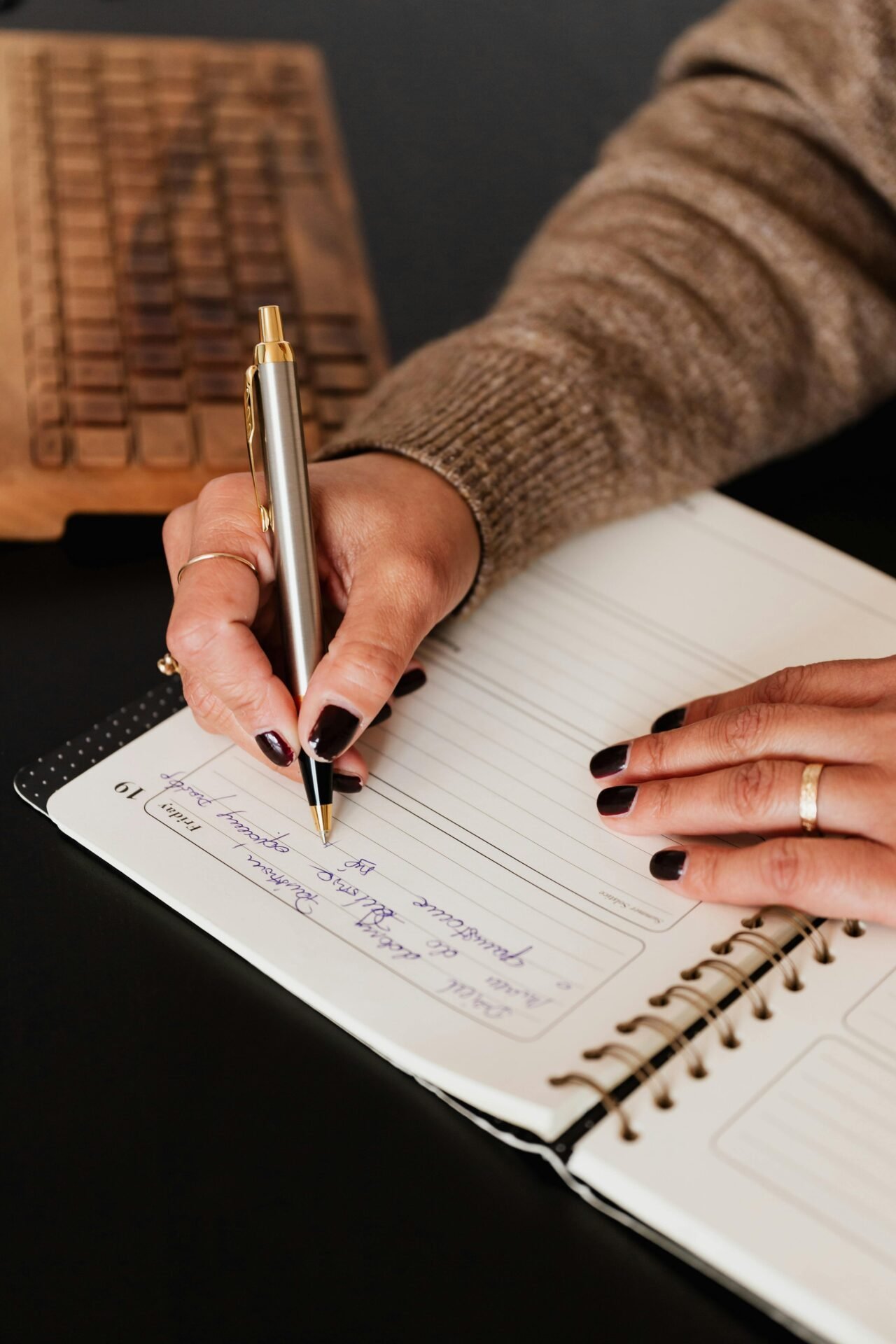 Eine Frau mit dunkelrotem Nagellack und einem braunen Wollpullover schreibt in ihren Kalender, als würde sie morgens ihren Tag planen.