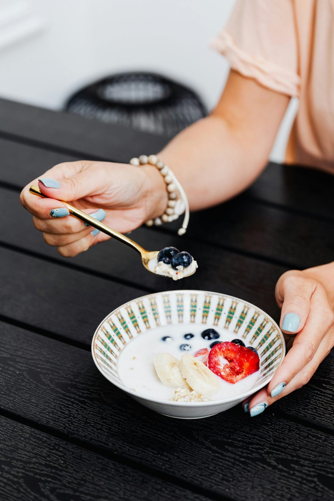 Es sind die Hände einer Frau zu sehen, die ein Armband trägt und hellblaue Fingernägel mit Blumen hat. Sie löffelt Porridge mit Erdbeeren, Bananen und Blaubeeren aus einer Porzellanschüssel, wahrscheinlich ein Frühstück.
