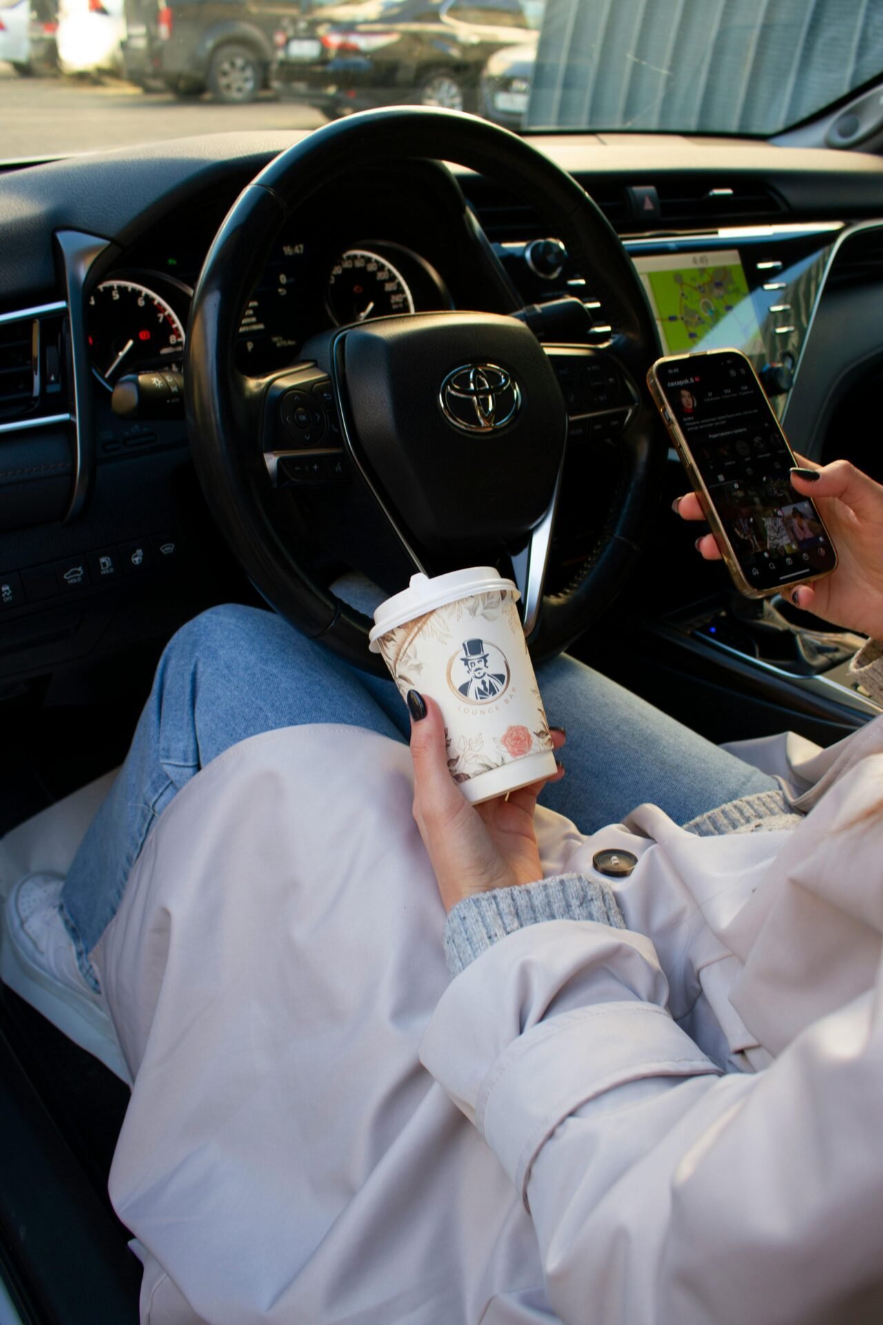Eine Frau sitzt in der Morgensonne in ihrem Auto, in einer Hand einen Kaffeebecher, in der anderen ihr Handy - eine Morgenroutine, die mit Pendeln vereinbar ist.