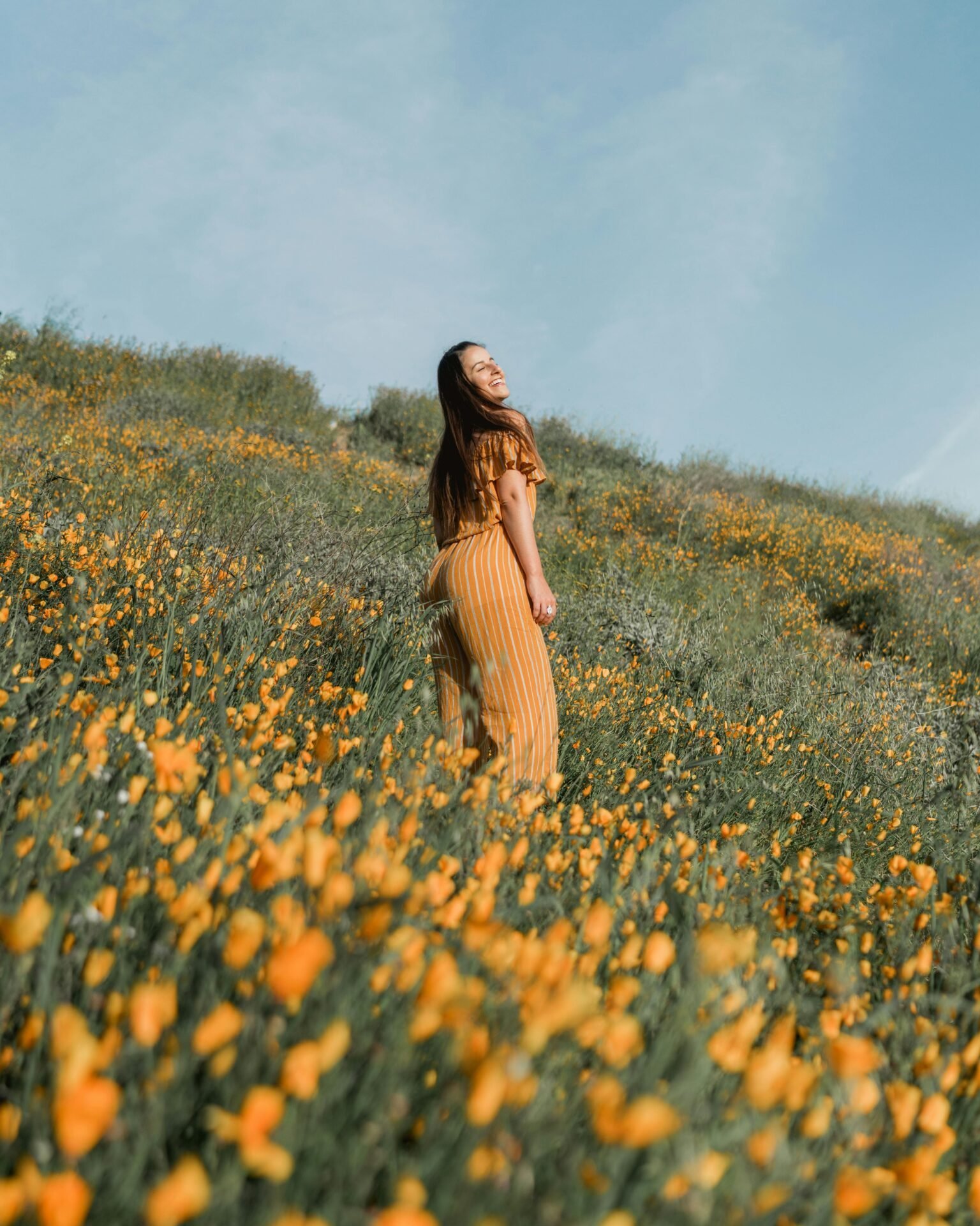 Eine junge Frau in einem gelben Jumpsuit genieße einen Glücksmoment auf einer Wiese mit gelben Blumen - sie lacht.