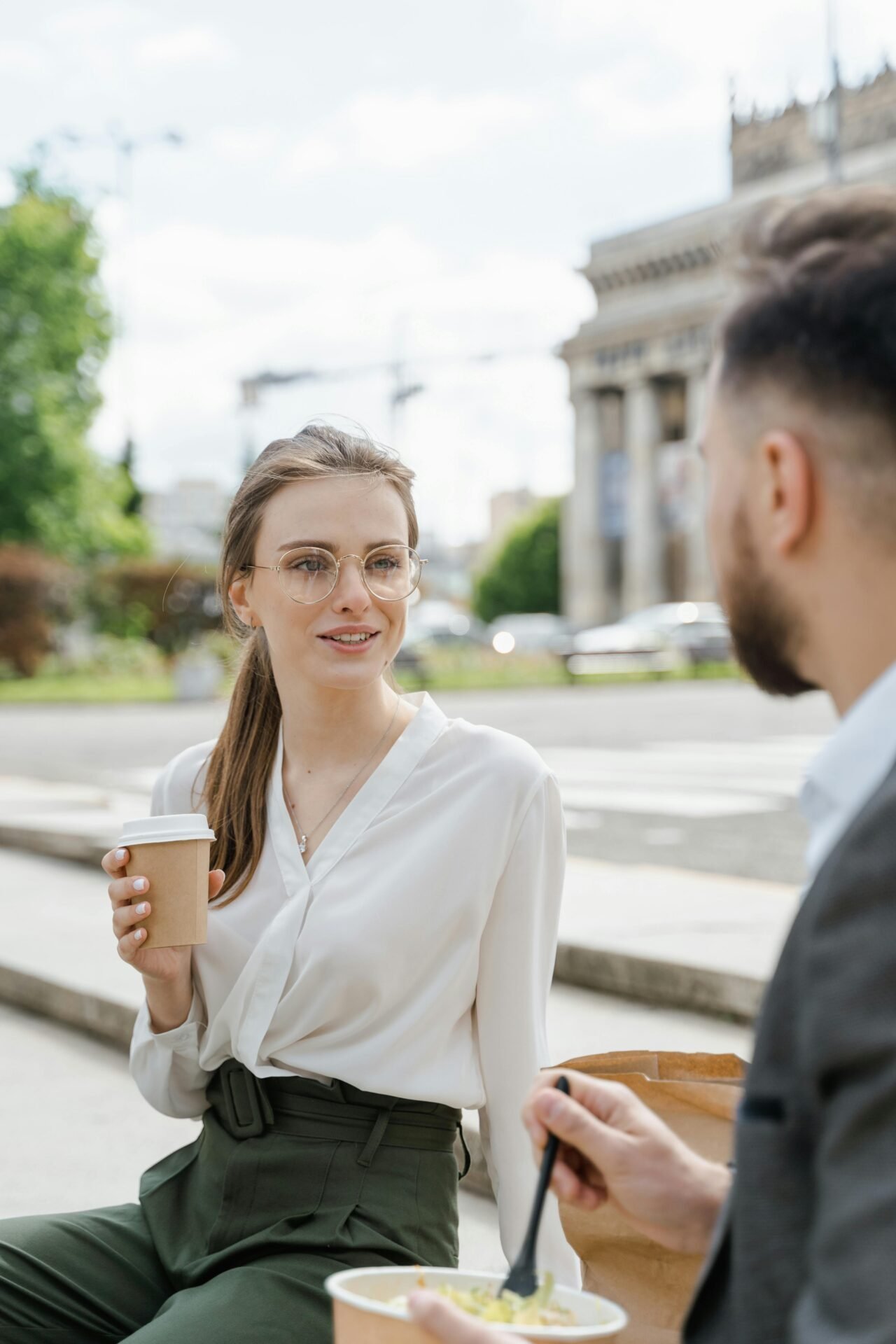 Eine Business-Frau mit einem Kaffee unterhält sich auf einer Bank mit einem Kollegen, der einen Salat isst.