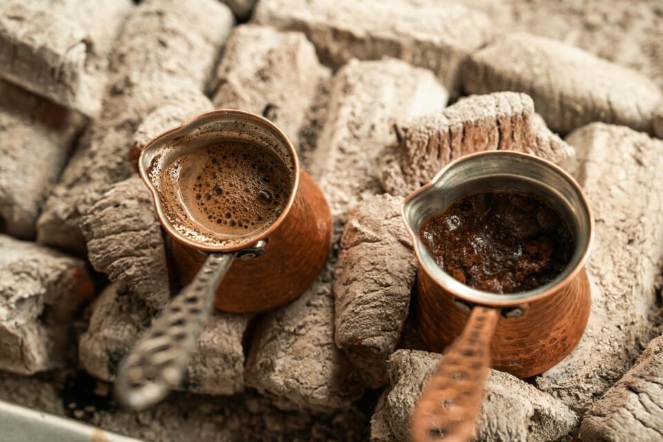 Zwei Kupferkannen mit Kaffee stehen auf heißen Kohlen. Der brodelnde Kaffee in jeder Kanne zeigt, dass sie erhitzt werden. Die Kannen haben lange, verzierte Griffe. Im Hintergrund befindet sich ein Bett aus aschebedeckter Kohle, was auf eine traditionelle Brühmethode hindeutet.