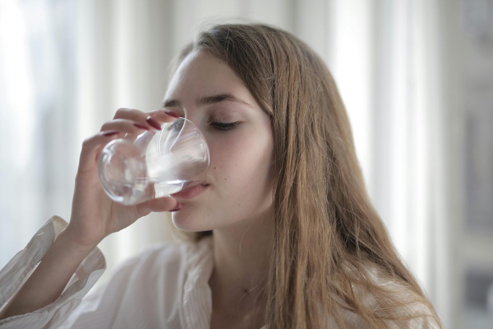 Eine Person mit langen Haaren und einem weißen Hemd trinkt Wasser aus einem durchsichtigen Glas. Sie befindet sich in einem Gebäude, in dem durch die großen Fenster weiches Tageslicht einfällt.