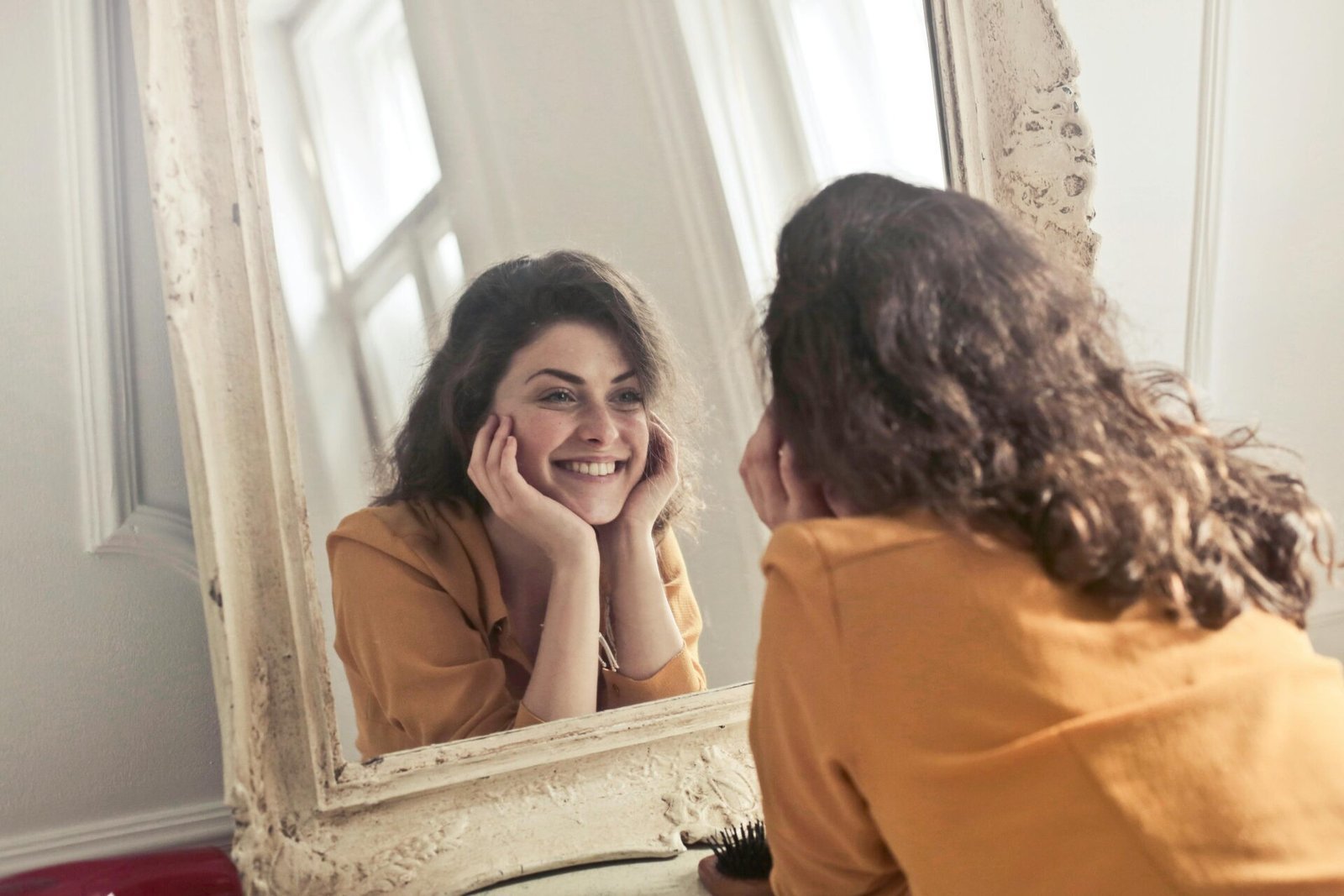 Eine Frau mit lockigem braunem Haar lächelt ihr Spiegelbild in einem Vintage-Spiegel an. Sie trägt ein senffarbenes Oberteil und stützt ihr Kinn auf ihre Hände. Sie strahlt ein fröhliches und zufriedenes Wesen aus.