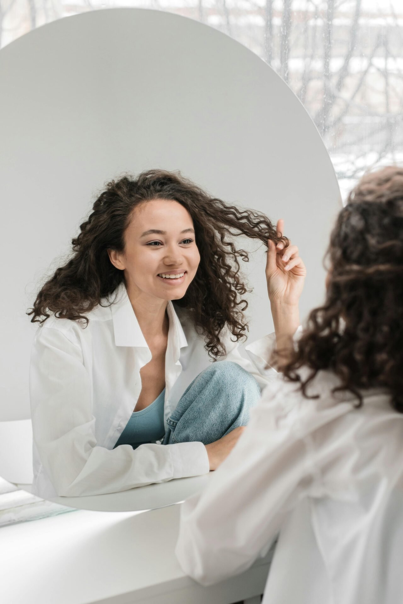 Eine Frau mit braunen Locken und einer weißen Bluse sieht sich lächelnd im Spiegel an.