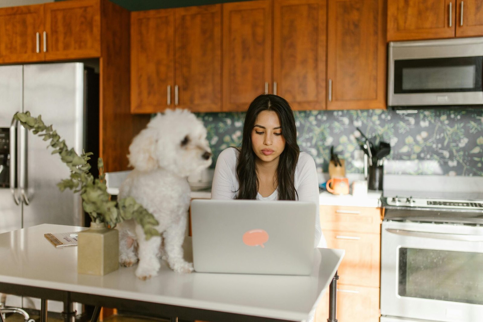 Eine Frau sitzt an einer Kücheninsel und benutzt einen Laptop. Neben ihr steht ein flauschiger weißer Hund. Die Küche ist mit Holzschränken und einer Küchenrückwand mit Blättermuster ausgestattet. Auf der Arbeitsplatte steht eine Vase mit getrocknetem Eukalyptus.
