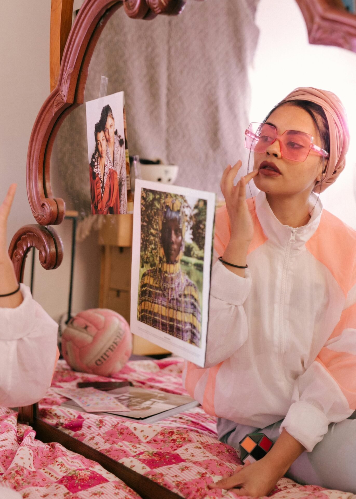 Person mit rosa Sonnenbrille, die eine starke Persönlichkeit ausstrahlt und auf einem Bett neben einem mit Fotos bedeckten Spiegel sitzt.