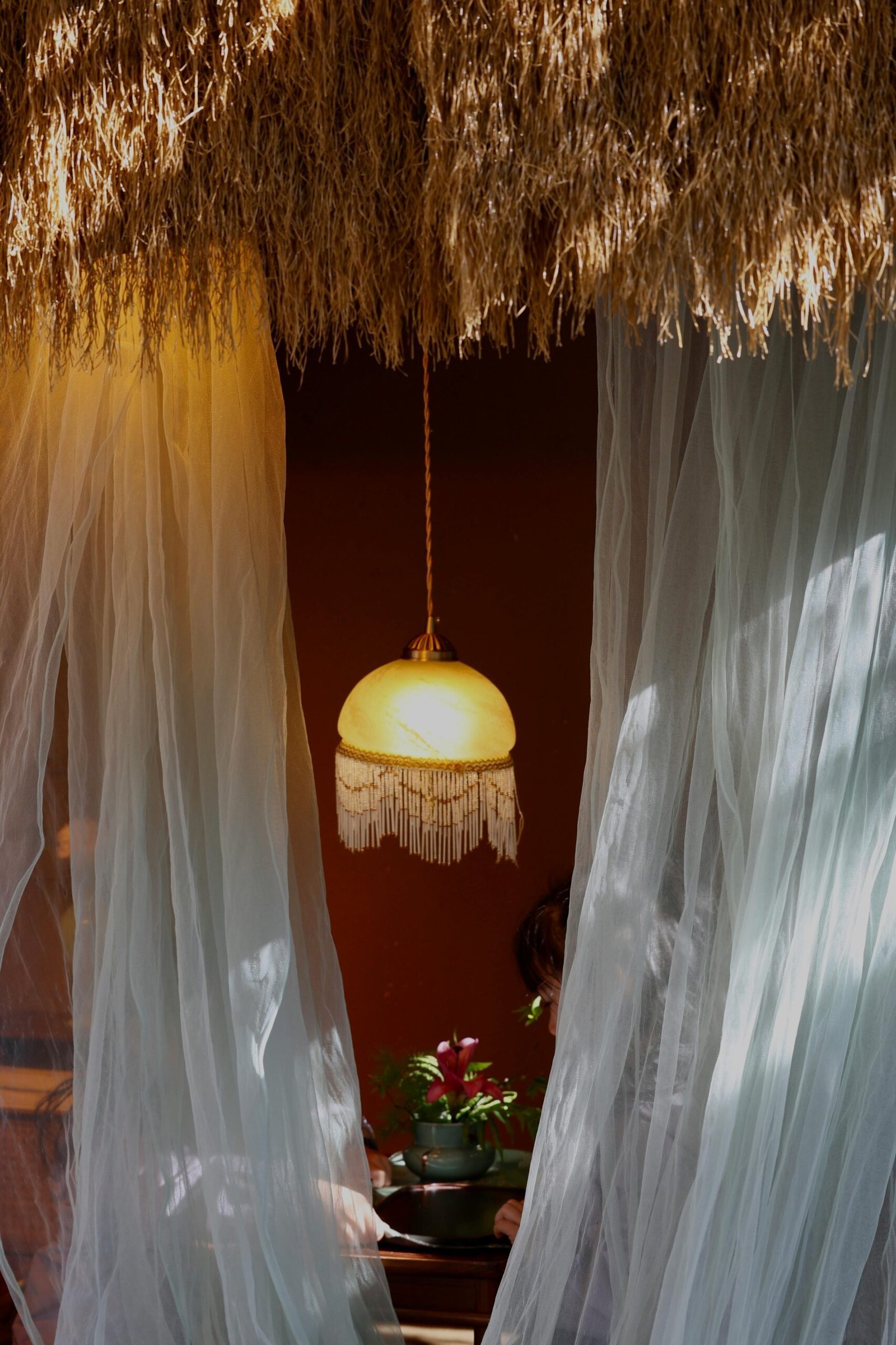 Eine orientalische Hängelampe strahlt sanftes Licht aus, während hinter einem weißen Vorhang eine Frau mit einer exotischen Blume sitzt – eine Atmosphäre der Ruhe und Reflexion.