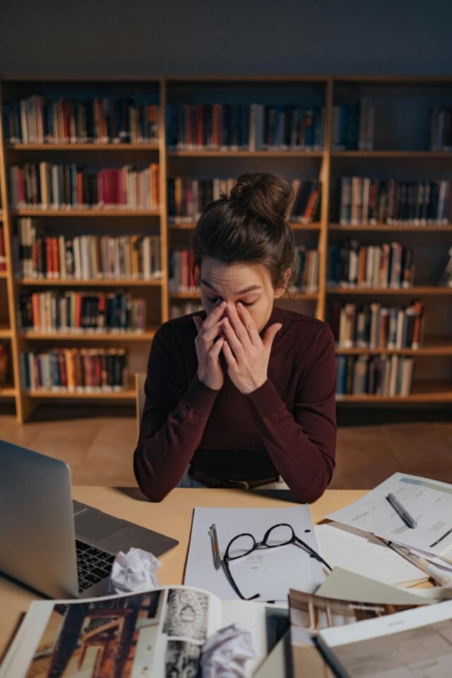 Person am Schreibtisch in der Bibliothek, reibt sich die Augen, umgeben von Büchern und Papieren – ein klassisches toxisches Arbeitsumfeld.