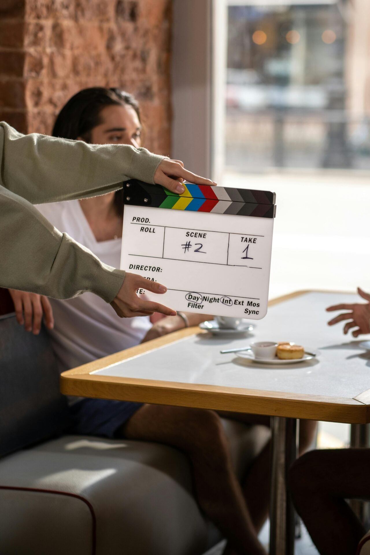 Eine Schauspielerin wie Jennifer Aniston sitzt verschwommen mit jemandem in einem Café und redet. Im Vordergrund sieht man eine Film-Klappe die vor den beiden gerade geschlossen wird.