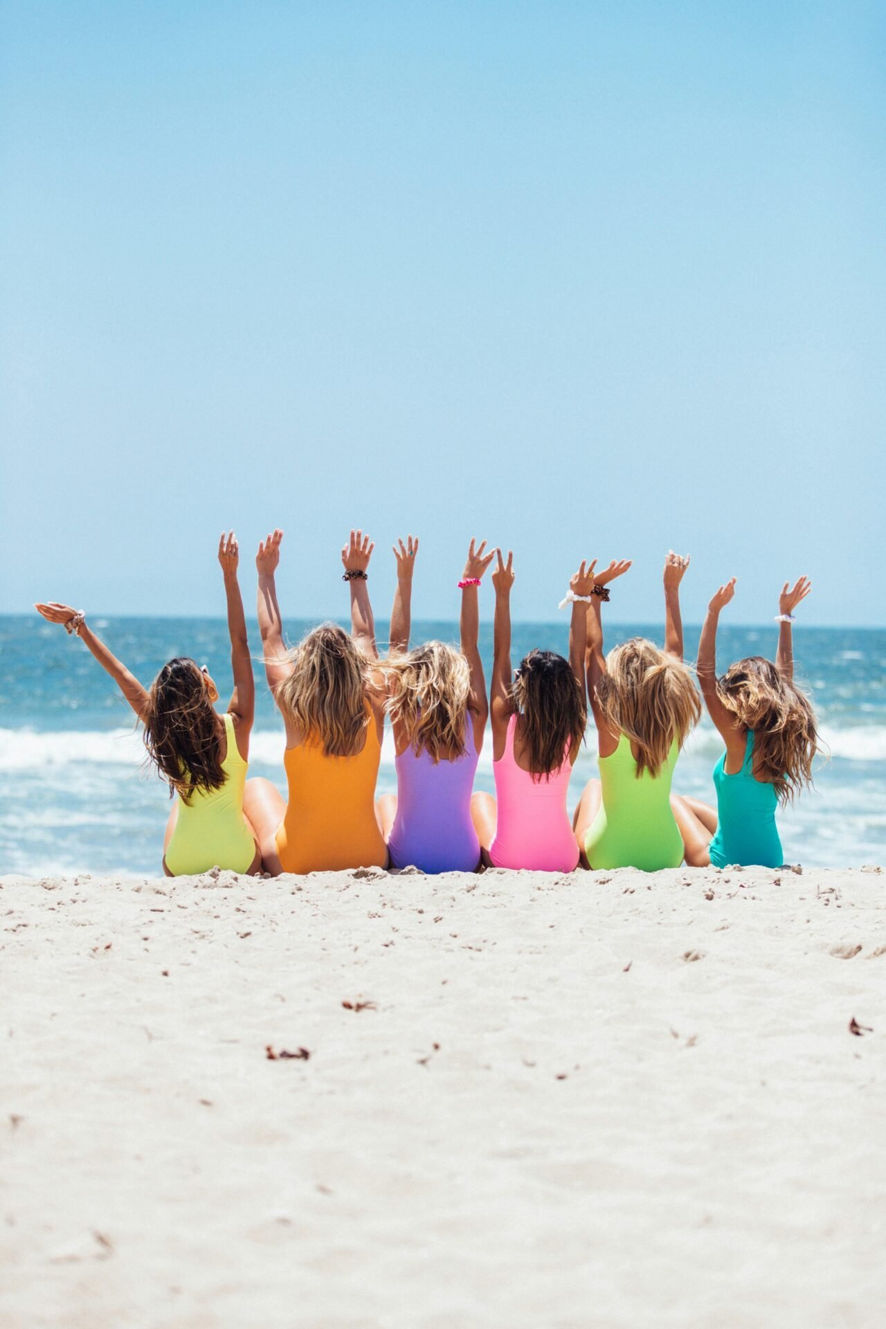 Sechs Menschen in bunten Badeanzügen sitzen mit erhobenen Armen an einem Strand und blicken auf das Meer.