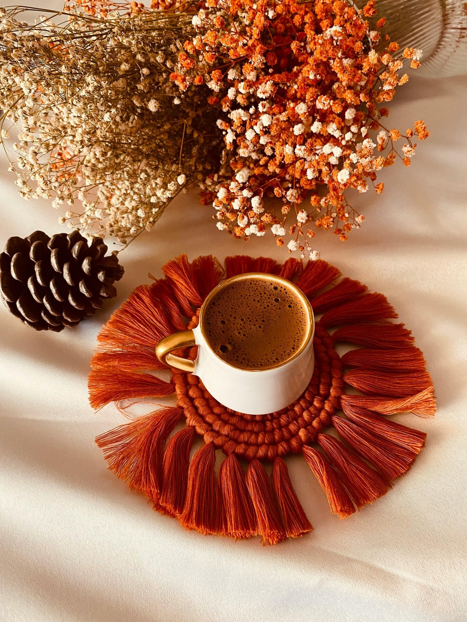 Eine Tasse Kaffee auf einem orangen Untersetzer, umgeben von getrockneten Blumen und einem Tannenzapfen – eine gemütliche Szene für eine kleine Auszeit.