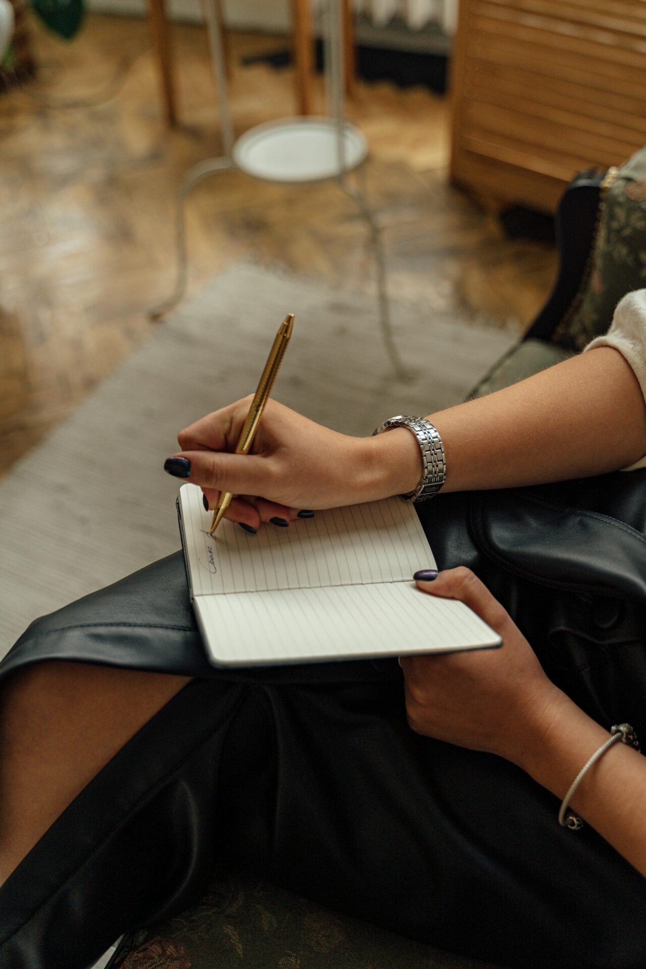 Eine Person mit dunklem Nagellack schreibt mit einem goldenen Stift in ein liniertes Notizbuch. Sie sitzt auf einem grünen Sofa und hat eine schwarze Ledertasche auf dem Schoß. Im Hintergrund sind ein Teppich und ein Teil einer Lampe zu sehen.