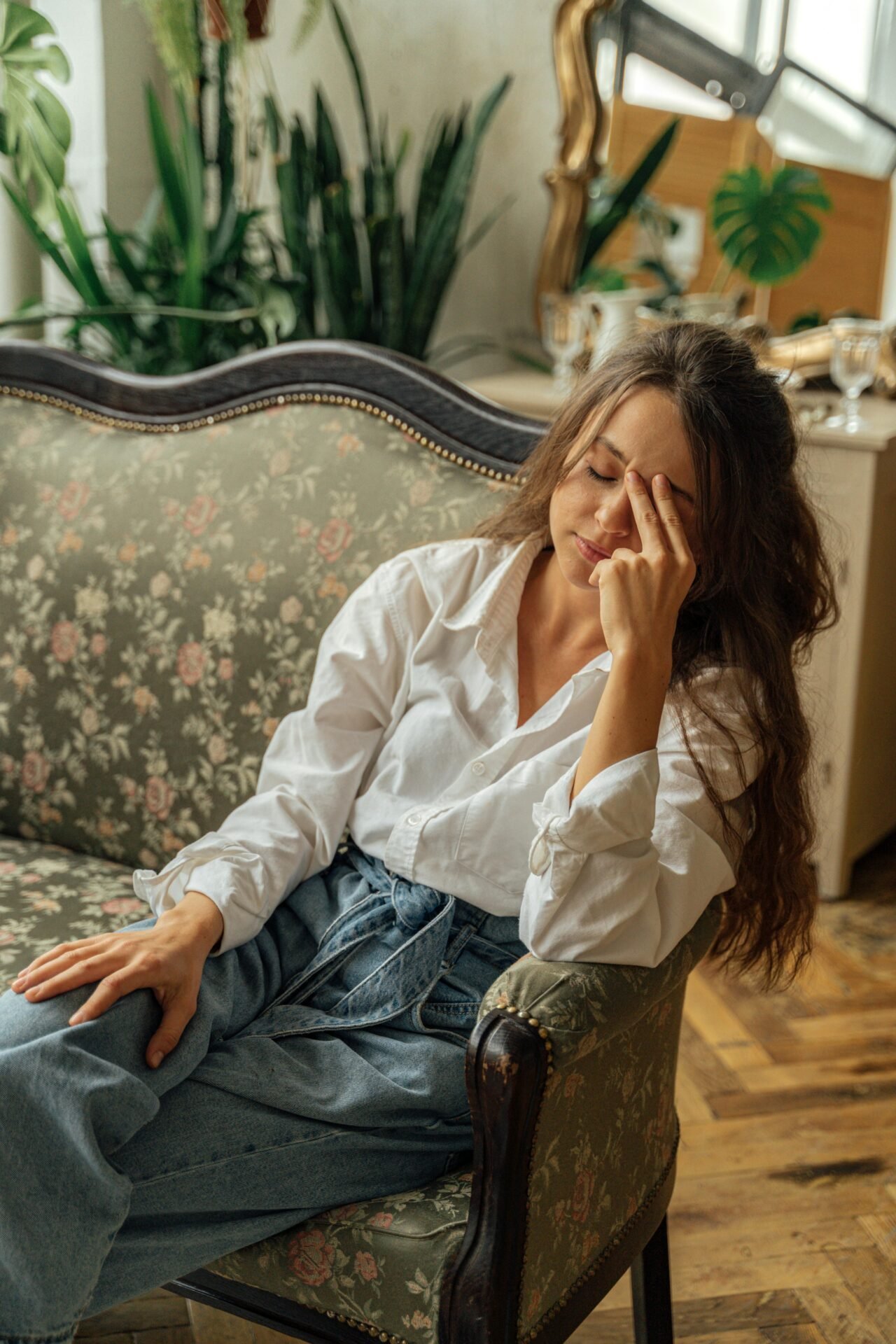 Frau in weißem Hemd und Jeans sitzt auf einem geblümten Sofa und lächelt sanft – ein Bild für gesunden Lebensstil.