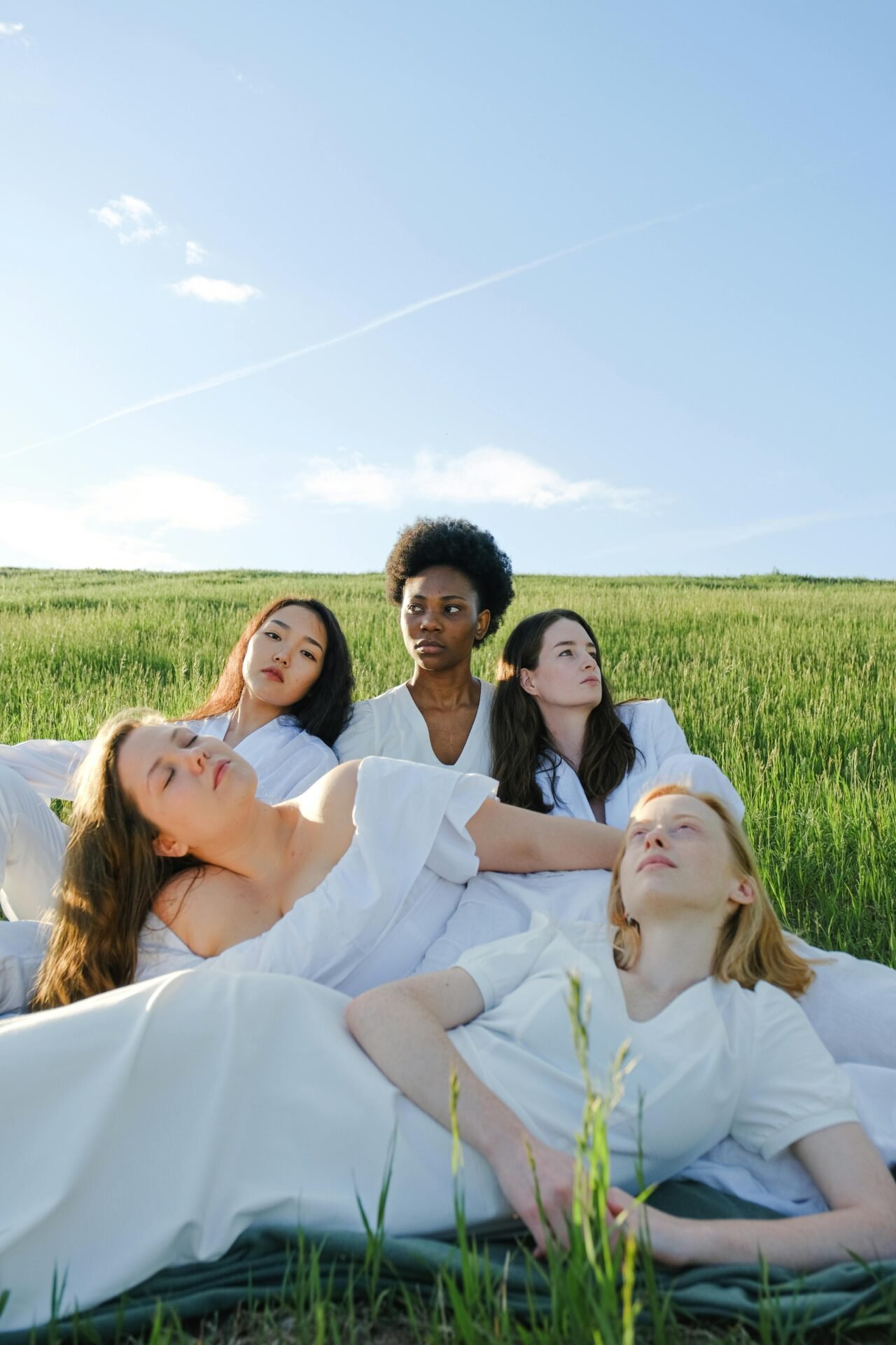 Fünf sehr unterschiedliche Frauen in weißer Kleidung entspannen sich auf einer Wiese und umarmen sich unter blauem Himmel.
