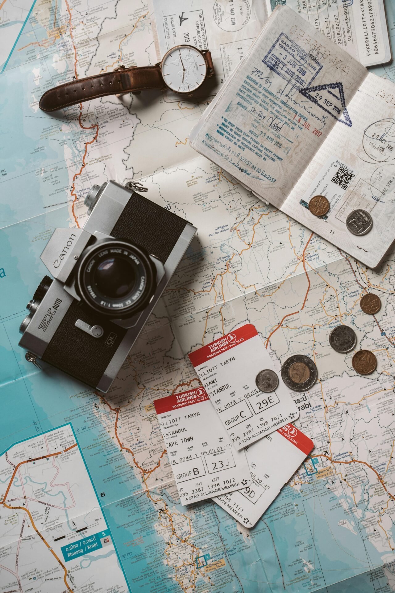 Flachgelegte Essentials für eine Reise ins Glück: Kamera, Karte, Reisepass, Uhr, Münzen, Bordkarten.