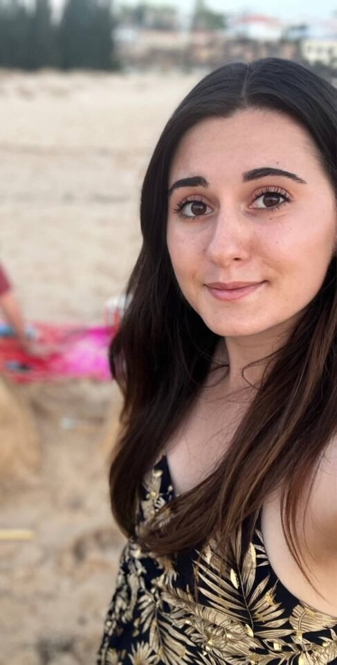 Suzan Serra, eine Frau mit langen dunklen Haaren und einem geblümten Kleid lächelt an einem Sandstrand in die Kamera. Im Hintergrund sind verschwommene Bilder von Strandstühlen und Bäumen zu sehen.