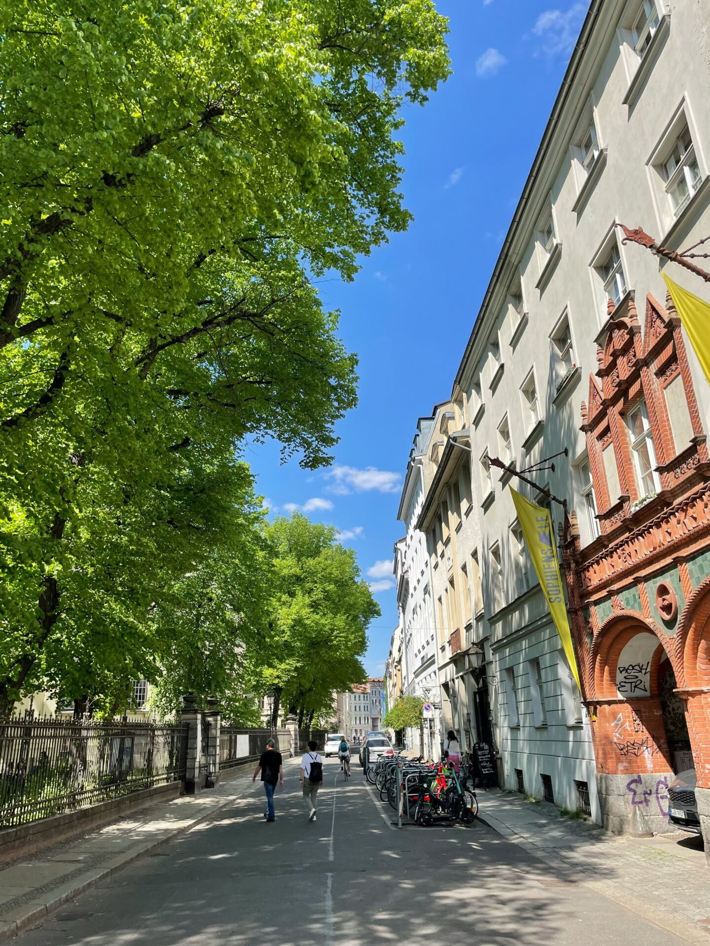 Menschen gehen eine sonnige Berliner Straße entlang, die von Bäumen und historischen Sehenswürdigkeiten gesäumt ist.