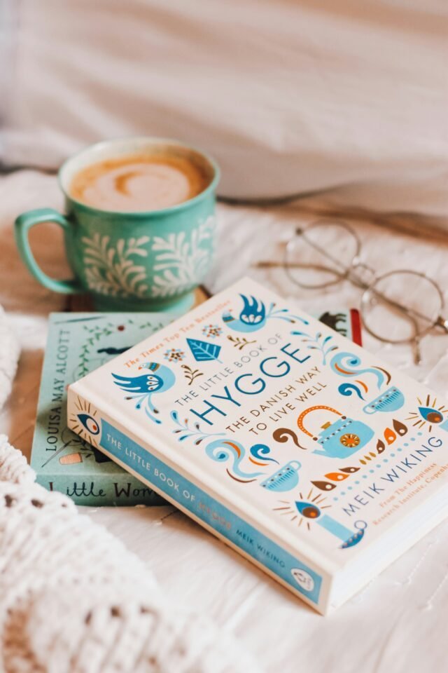 Eine gemütliche Szene mit „The Little Book of Hygge“ auf „Little Women“ von Louisa May Alcott, platziert auf einem Bett. Daneben stehen ein paar Gläser und eine Tasse mit einem heißen Getränk, alles umgeben von weicher weißer Bettwäsche.